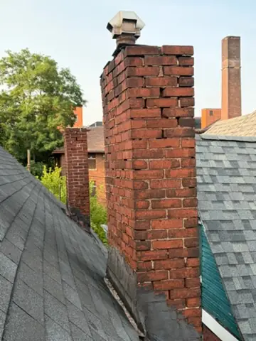 Tall older red brick chimney with a metal cap on a Pittsburgh house, an industrial flue stack visible behind it