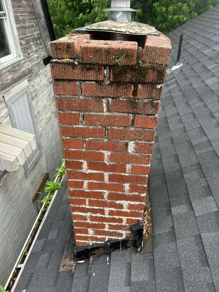 Red brick chimney with washed-out mortar joints and torn base flashing before tuckpointing in Brackenridge, PA