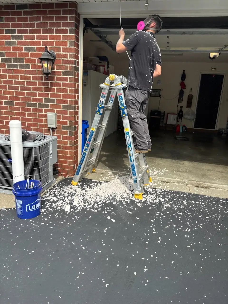 The Chimney Experts technician on a ladder in a respirator, feeding a cleaning rod into an overhead dryer vent line in a Murrysville, PA garage